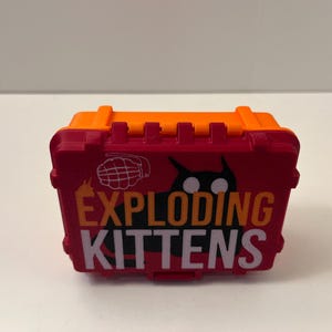 K&ouml;nnte beinhalten: Eine rot-orange Exploding Kittens Spielbox. Die Box zeigt das Logo des Spiels in Wei&szlig; und Orange, eine schwarze Katzen-Silhouette und eine Granaten-Grafik. Die Box ist aus Kunststoff und hat einen Griff oben.