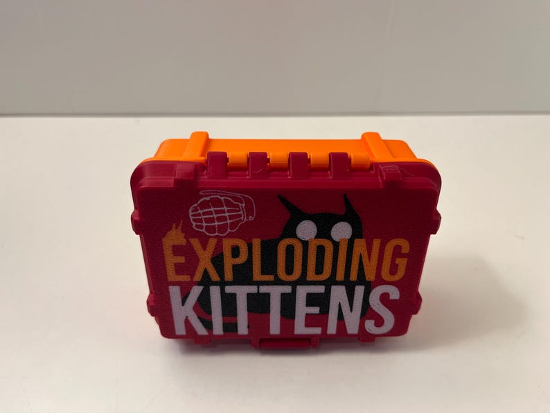 K&ouml;nnte beinhalten: Eine rot-orange Exploding Kittens Spielbox. Die Box zeigt das Logo des Spiels in Wei&szlig; und Orange, eine schwarze Katzen-Silhouette und eine Granaten-Grafik. Die Box ist aus Kunststoff und hat einen Griff oben.