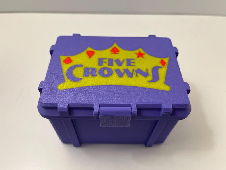 May include: A purple plastic box featuring the text "FIVE CROWNS" in yellow, with a crown, heart, diamond, spade, and star graphic. The box has a hinged lid and a latch. The box is designed for storing game pieces.