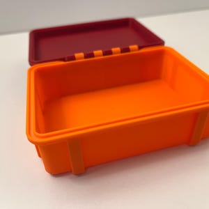 K&ouml;nnte beinhalten: Ein offener, rechteckiger Beh&auml;lter in Orange mit einem weinroten Deckel. Der Beh&auml;lter hat eine glatte Oberfl&auml;che und ist mit kleinen rechteckigen St&uuml;tzen an den Seiten versehen. Der Deckel ist leicht ge&ouml;ffnet und gibt den Blick ins Innere frei.