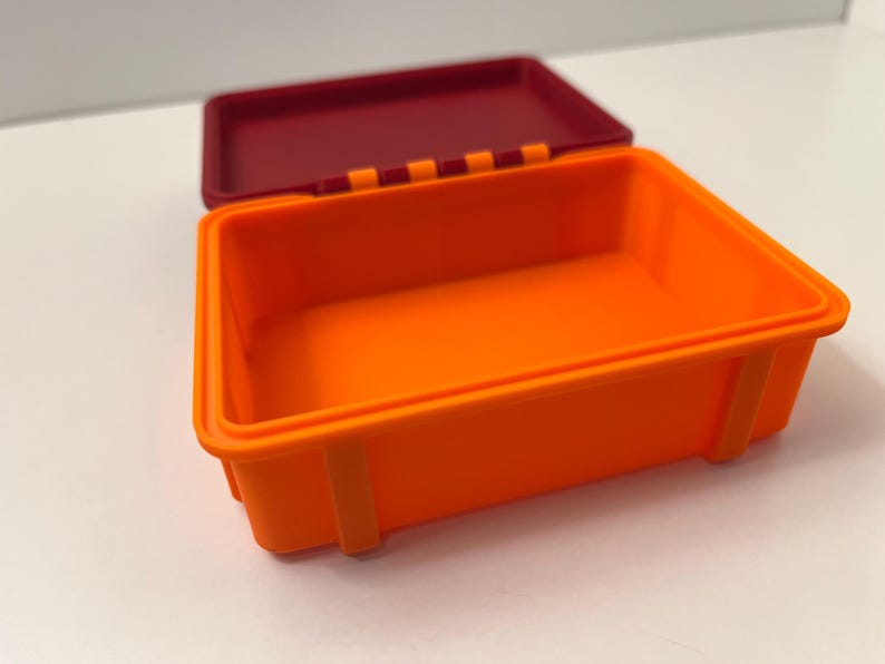 K&ouml;nnte beinhalten: Ein offener, rechteckiger Beh&auml;lter in Orange mit einem weinroten Deckel. Der Beh&auml;lter hat eine glatte Oberfl&auml;che und ist mit kleinen rechteckigen St&uuml;tzen an den Seiten versehen. Der Deckel ist leicht ge&ouml;ffnet und gibt den Blick ins Innere frei.