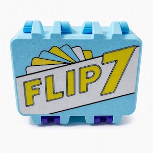May include: A light blue rectangular storage case with a textured surface and purple hinges. The front features a white diagonal banner with the yellow text FLIP 7 and a graphic of fanned-out cards in yellow, white, and blue colors.