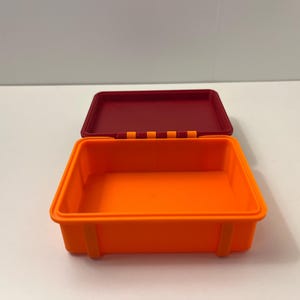 K&ouml;nnte beinhalten: Ein offener, rechteckiger, orangefarbener Beh&auml;lter mit einem weinroten Deckel. Der Beh&auml;lter hat eine glatte Oberfl&auml;che und abgerundete Ecken. Der Deckel ist leicht ge&ouml;ffnet und gibt den Blick ins Innere frei. Der Beh&auml;lter steht auf einer wei&szlig;en Oberfl&auml;che.