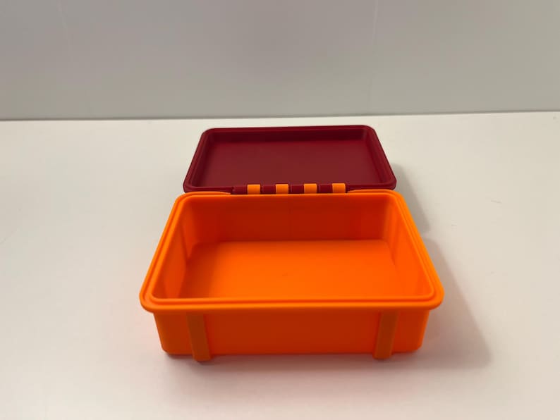 K&ouml;nnte beinhalten: Ein offener, rechteckiger, orangefarbener Beh&auml;lter mit einem weinroten Deckel. Der Beh&auml;lter hat eine glatte Oberfl&auml;che und abgerundete Ecken. Der Deckel ist leicht ge&ouml;ffnet und gibt den Blick ins Innere frei. Der Beh&auml;lter steht auf einer wei&szlig;en Oberfl&auml;che.