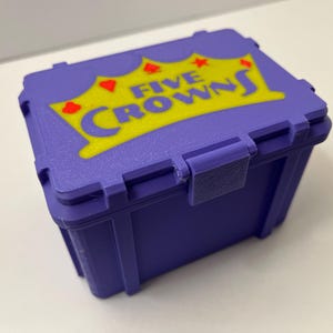 May include: A purple plastic box featuring the text "FIVE CROWNS" in yellow and red. The box has a hinged lid and a clasp. This box is designed to store a card game.