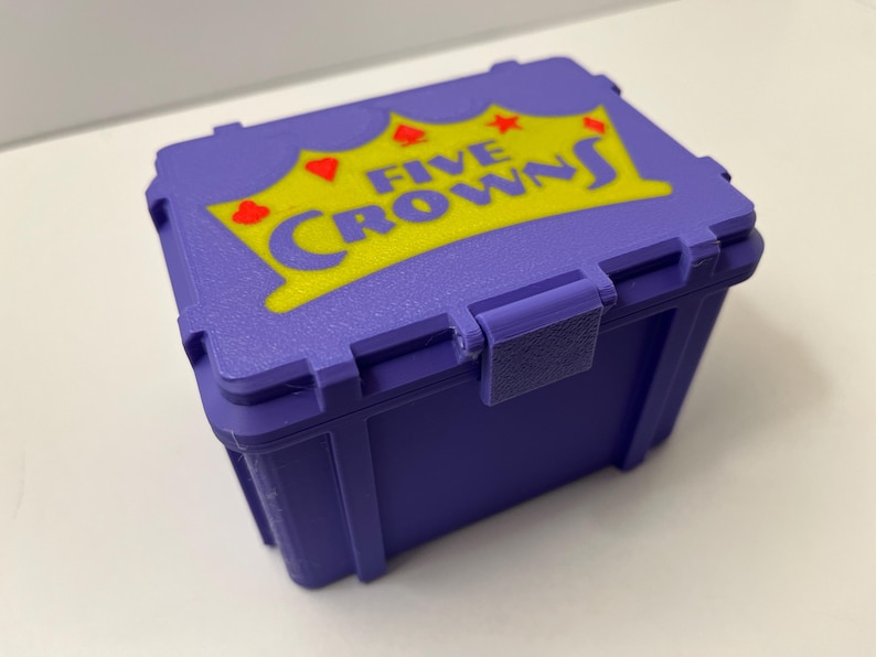 May include: A purple plastic box featuring the text "FIVE CROWNS" in yellow and red. The box has a hinged lid and a clasp. This box is designed to store a card game.