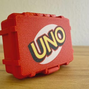 Puede incluir: Una caja roja texturizada con la palabra "UNO" en letras amarillas grandes sobre un óvalo blanco. La caja tiene forma rectangular con bordes redondeados y un asa pequeña. La caja del juego está sobre una superficie de madera.