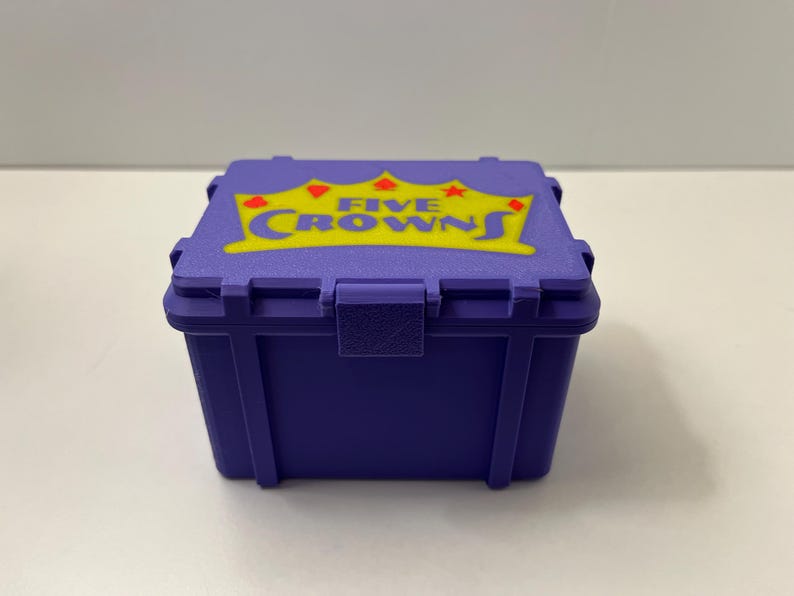 May include: A small, purple box with a hinged lid. The lid displays the words "FIVE CROWNS" in yellow, with a crown and star design. The box is made of a durable material and has a secure latch.