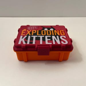 K&ouml;nnte beinhalten: Eine rechteckige, zweifarbige Box mit den Worten "EXPLODING KITTENS" in wei&szlig;er und orangefarbener Schrift. Der Deckel ist rot mit einer Grafik einer Katze und eines Globus. Die Basis ist orange.