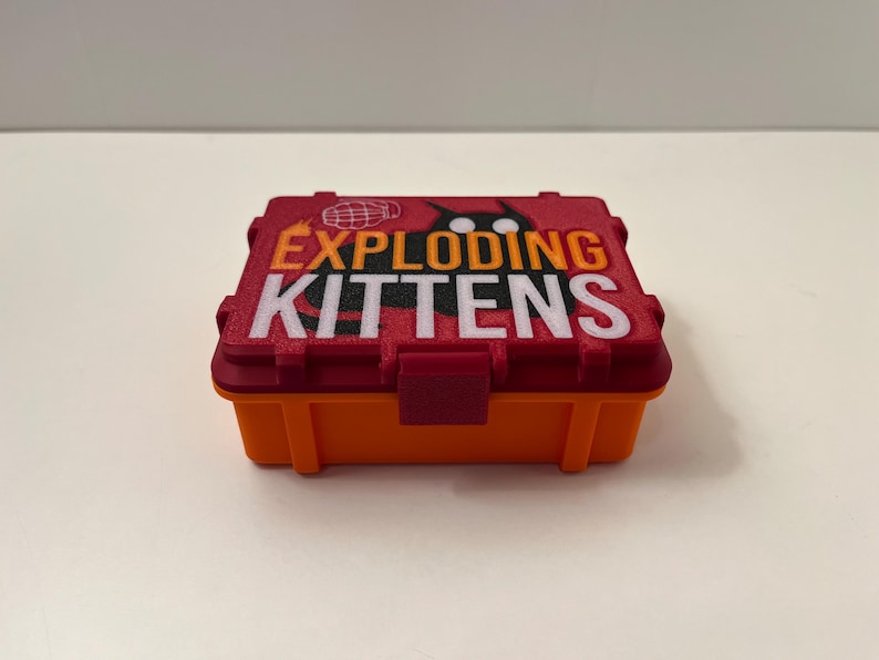 K&ouml;nnte beinhalten: Eine rechteckige, zweifarbige Box mit den Worten "EXPLODING KITTENS" in wei&szlig;er und orangefarbener Schrift. Der Deckel ist rot mit einer Grafik einer Katze und eines Globus. Die Basis ist orange.