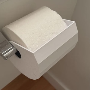May include: A white toilet paper holder with a geometric, faceted design mounted on a wall. The holder features a chrome-finished metal arm that attaches to the wall, supporting a roll of white toilet paper within the angular white plastic frame.