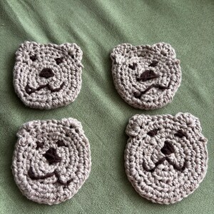 May include: Four handmade crochet coasters in the shape of a bear's face. The coasters are light gray with dark brown facial features, including the nose and mouth. The coasters are arranged on a green surface.