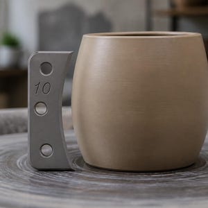 May include: A smooth, light brown ceramic mug with a rounded body shape. Positioned next to the mug is a grey, vertical pottery rib tool featuring three circular cutouts and the number 10 embossed on its surface, used for shaping clay.