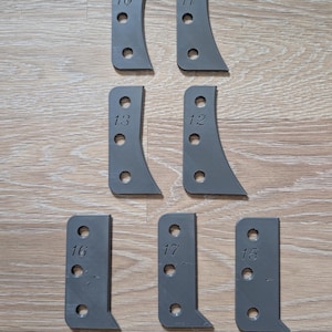 Könnte beinhalten: Acht graue Metallhalterungen in verschiedenen Formen und Größen. Jede Halterung hat vorgebohrte Löcher und ist mit einer Nummer von 10 bis 17 versehen. Die Halterungen sind auf einer hellbraunen Holzoberfläche angeordnet.
