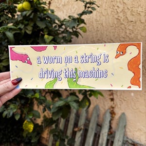 Puede incluir: Una pegatina rectangular con el texto "a worm on a string is driving this machine" en morado y blanco. La pegatina presenta gusanos de dibujos animados en rosa, verde y naranja sobre un fondo amarillo claro con confeti colorido.