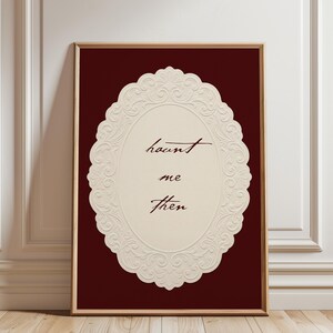 May include: Framed art print featuring the words "haunt me then" in elegant script within a cream-colored, ornate oval design. The background is a deep burgundy, and the frame is a warm brown.