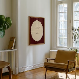 May include: A framed artwork with the text "haunt me then" in a decorative oval shape. The artwork is set against a burgundy background and framed in wood. A mid-century modern chair and a copy of "Great Gatsby" are also visible.