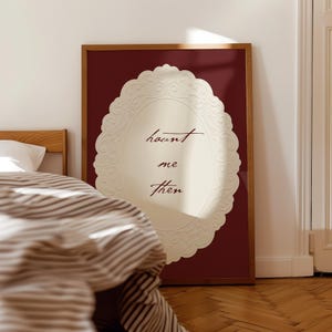 May include: A framed wall art piece with a burgundy background and a cream-colored oval design. The oval has a decorative, embossed border and the words "haunt me then" in a handwritten font. The frame is a warm brown color.