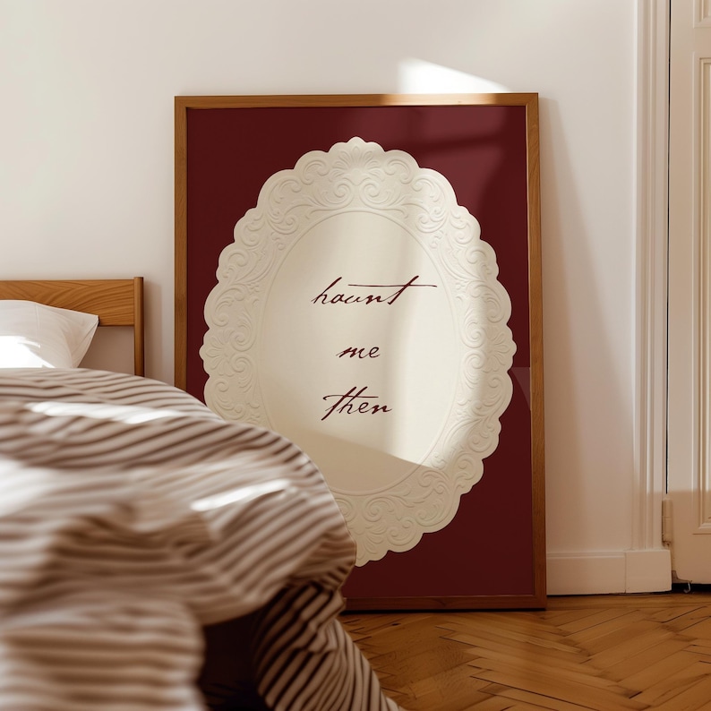 May include: A framed wall art piece with a burgundy background and a cream-colored oval design. The oval has a decorative, embossed border and the words "haunt me then" in a handwritten font. The frame is a warm brown color.