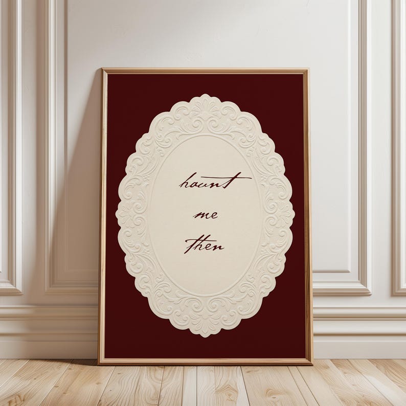 May include: Framed art print featuring the words "haunt me then" in elegant script within a cream-colored, ornate oval design. The background is a deep burgundy, and the frame is a warm brown.