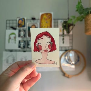 May include: A small rectangular watercolor painting on paper featuring a stylized portrait of a person with short red hair, pink cheeks, and dark ink outlines. The artwork includes a signature in the bottom right corner with the letters ef.