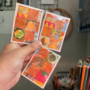 May include: Three rectangular cards encased in clear plastic sleeves, each featuring a vibrant collage of orange, yellow, and green paper scraps, geometric patterns, and floral motifs, accented with circular metallic copper-colored stickers.