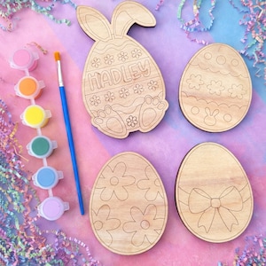 May include: Wooden Easter egg and bunny cutouts with floral and bow designs, along with a paint set and brush. The bunny cutout is personalized with the name "Hadley". The background is pink and purple with colorful paper shreds. Option 1 is written in white text.