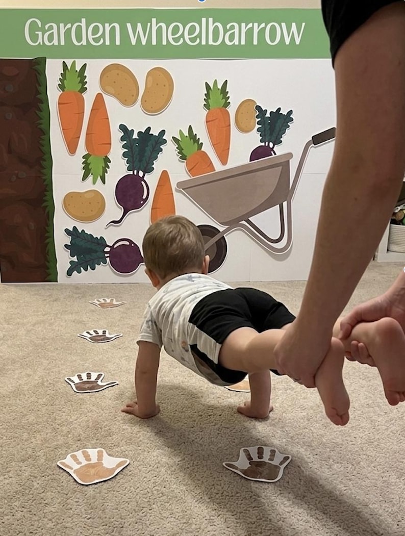 May include: A child crawling towards a garden-themed backdrop featuring carrots, potatoes, beets, and a wheelbarrow. The words "Garden wheelbarrow" are at the top. Footprint cutouts are on the carpet.