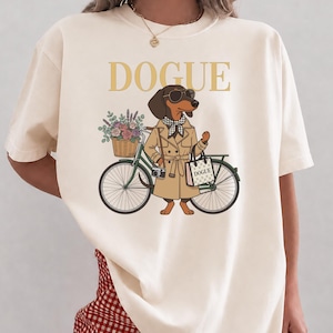 May include: An ivory t-shirt featuring a graphic of a dachshund wearing a tan trench coat, a checkered scarf, and sunglasses. The dog stands next to a green bicycle with a basket of flowers and carries a shopping bag labeled DOGUE.