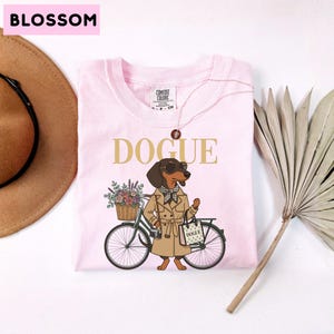 May include: A light pink t-shirt featuring a graphic of a dachshund wearing a tan trench coat, sunglasses, and a checkered scarf. The dog stands next to a bicycle with a flower basket and carries a shopping bag labeled DOGUE. The text DOGUE is printed above the dog in gold letters.