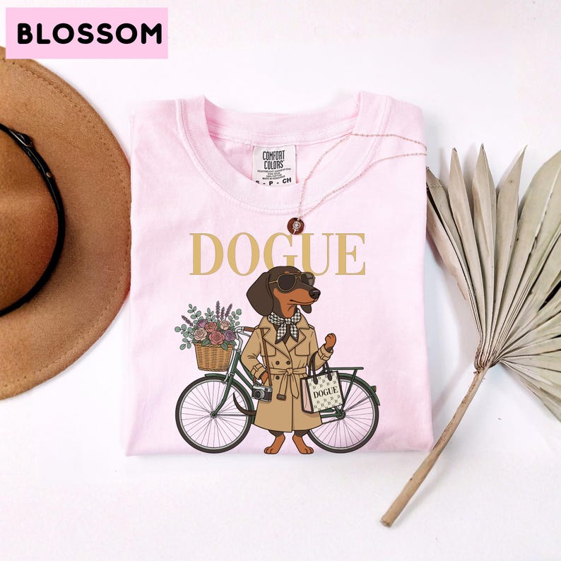 May include: A light pink t-shirt featuring a graphic of a dachshund wearing a tan trench coat, sunglasses, and a checkered scarf. The dog stands next to a bicycle with a flower basket and carries a shopping bag labeled DOGUE. The text DOGUE is printed above the dog in gold letters.