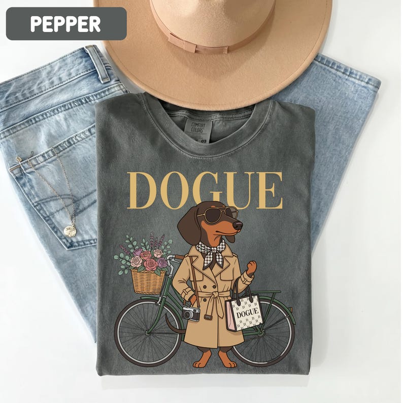 May include: A charcoal gray t-shirt featuring a graphic of a dachshund wearing a tan trench coat, sunglasses, and a checkered scarf. The dog stands next to a green bicycle with a flower basket and carries a shopping bag labeled DOGUE.