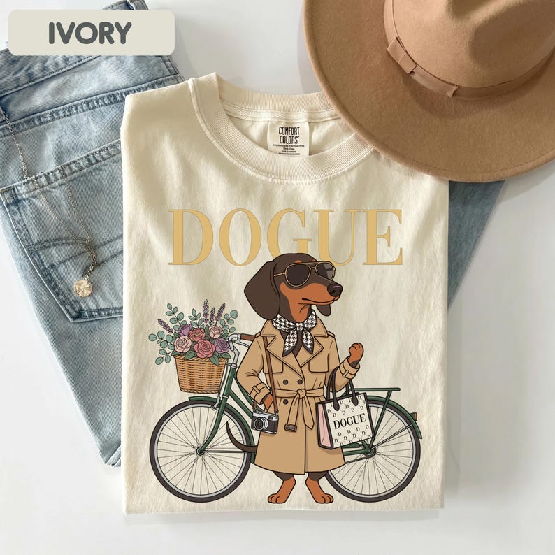 May include: An ivory t-shirt featuring a graphic of a dachshund wearing a tan trench coat, a checkered scarf, and sunglasses. The dog stands next to a green bicycle with a flower-filled basket and a tote bag labeled DOGUE. The text DOGUE is printed above.