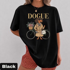 May include: A black t-shirt featuring a graphic of a dachshund wearing a tan trench coat, sunglasses, and a checkered scarf. The dog stands next to a bicycle with a flower basket, a camera, and a tote bag labeled DOGUE. The text DOGUE is printed above.