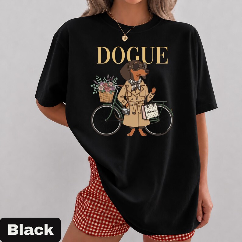 May include: A black t-shirt featuring a graphic of a dachshund wearing a tan trench coat, sunglasses, and a checkered scarf. The dog stands next to a bicycle with a flower basket, a camera, and a tote bag labeled DOGUE. The text DOGUE is printed above.