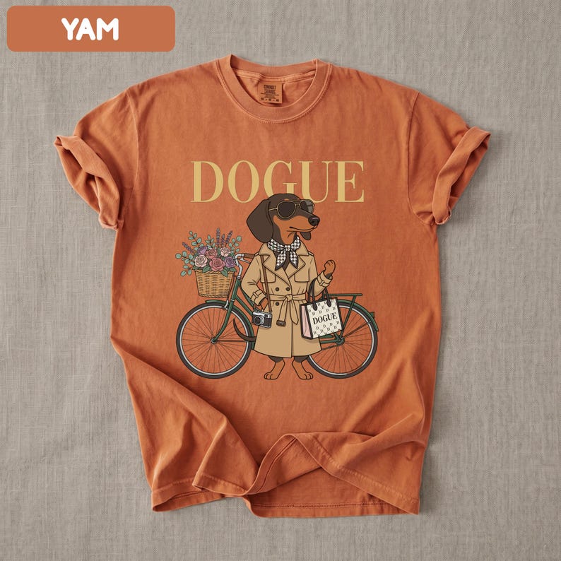 May include: A burnt orange t-shirt featuring a graphic of a dachshund wearing a tan trench coat, sunglasses, and a checkered scarf. The dog stands next to a bicycle with a flower basket and carries a shopping bag labeled DOGUE. The text DOGUE is printed above.
