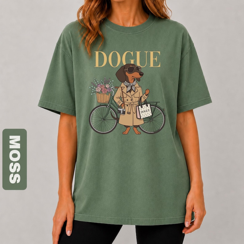 May include: A moss green short-sleeve t-shirt featuring a graphic of a dachshund wearing a tan trench coat, sunglasses, and a scarf. The dog stands next to a bicycle with a flower basket and carries a bag labeled DOGUE. The text DOGUE is printed above.
