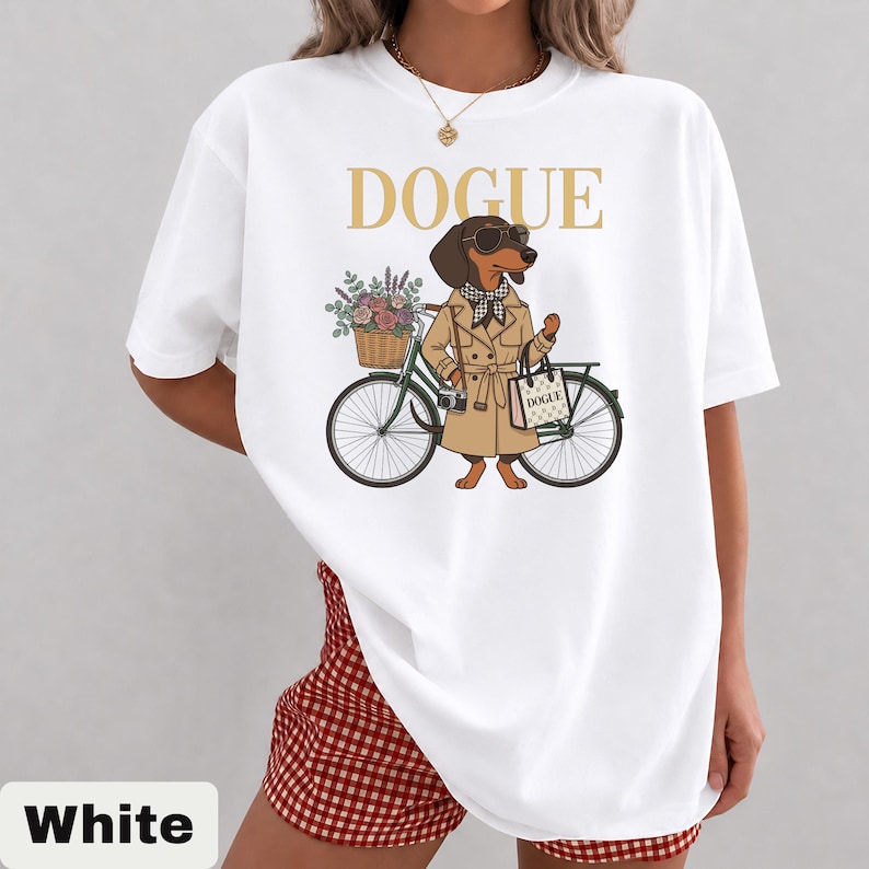 May include: A white t-shirt featuring a graphic of a brown dachshund wearing a tan trench coat, sunglasses, and a checkered scarf. The dog stands next to a bicycle with a flower basket and carries a shopping bag labeled DOGUE in gold text.