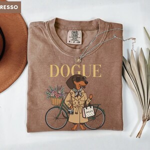 May include: An espresso-colored t-shirt featuring a graphic of a dachshund wearing a trench coat, sunglasses, and a checkered scarf. The dog stands next to a bicycle with a flower basket and carries a shopping bag labeled DOGUE.