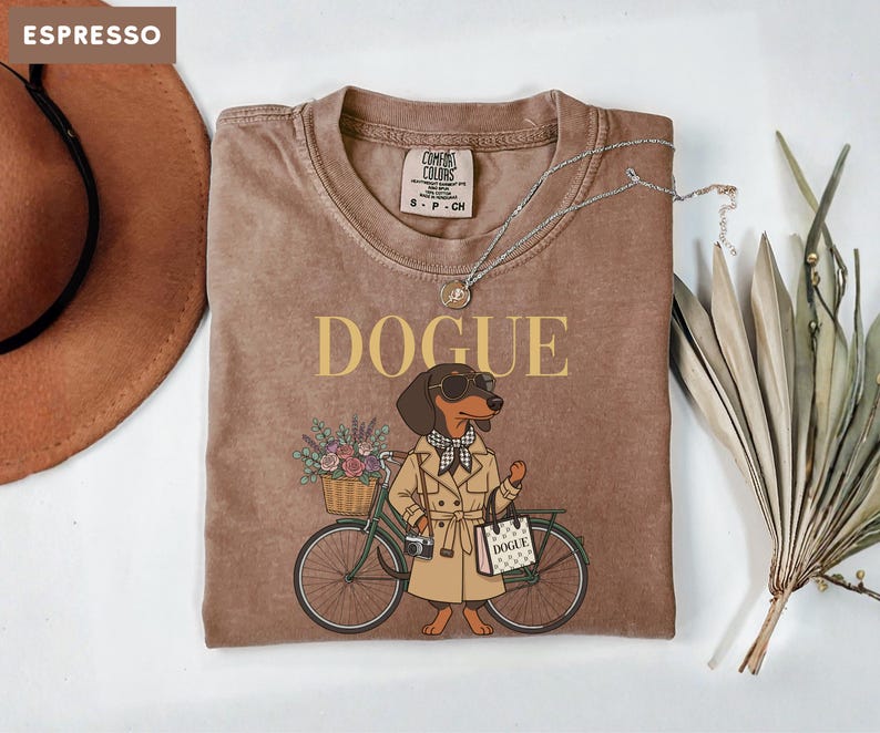 May include: An espresso-colored t-shirt featuring a graphic of a dachshund wearing a trench coat, sunglasses, and a checkered scarf. The dog stands next to a bicycle with a flower basket and carries a shopping bag labeled DOGUE.