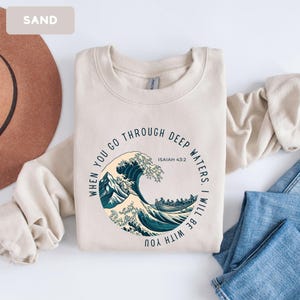 May include: Sand-colored sweatshirt with a circular graphic featuring a stylized wave, boat, and mountain scene. Text reads "WHEN YOU GO THROUGH DEEP WATERS. I WILL BE WITH YOU" and "ISAIAH 43:2".