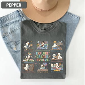 May include: A charcoal gray t-shirt featuring a graphic print of ducks engaged in various creative activities. The text reads "EXPLORE, CREATE, EVOLVE" along with captions like "Plant the seeds," "Navigate new horizons," "Construct your vision," "Find your flow," "Share your knowledge," "Examine detailed patterns," "Fuel your creativity," and "Rise to creative heights."