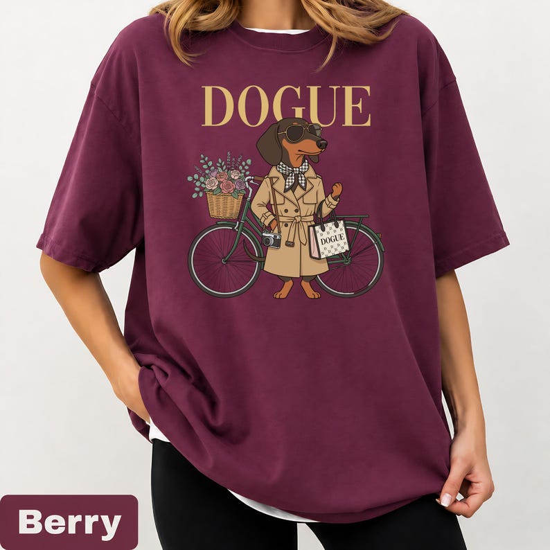 May include: A berry-colored t-shirt featuring a graphic of a dachshund wearing sunglasses, a checkered scarf, and a tan trench coat. The dog stands next to a bicycle with a flower-filled basket and carries a bag labeled DOGUE, with the text DOGUE above.