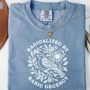 May include: Ice blue t-shirt with the words "RADICALIZED BY BASIC DECENCY" in white, featuring a bird and floral design. A gold necklace with a pendant rests on the shirt. The shirt has a Comfort Colors tag.