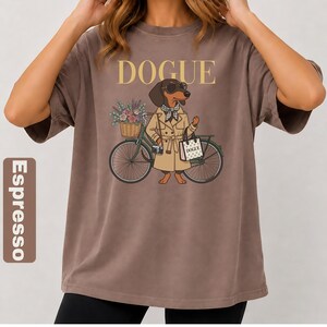 May include: An espresso-colored t-shirt featuring a graphic of a dachshund wearing a tan trench coat, sunglasses, and a checkered scarf. The dog stands next to a green bicycle with a flower basket and carries a bag labeled DOGUE.