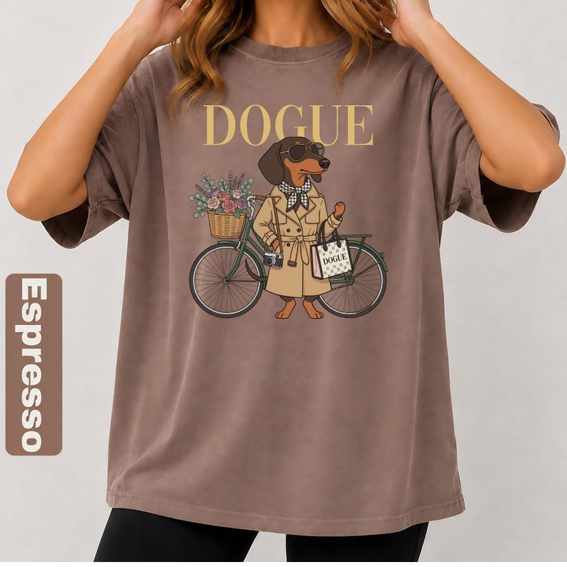 May include: An espresso-colored t-shirt featuring a graphic of a dachshund wearing a tan trench coat, sunglasses, and a checkered scarf. The dog stands next to a green bicycle with a flower basket and carries a bag labeled DOGUE.