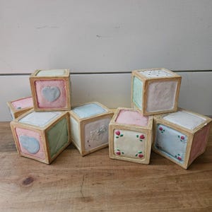 Może przedstawiać: Kolekcja sześciu dekoracyjnych kwadratowych klocków z jasnymi drewnianymi ramami. Każdy klocek posiada panele w pastelowych odcieniach różu, błękitu i bieli, ozdobione tłoczonymi wzorami, w tym sercami, drobnymi motywami kwiatowymi i kropkami.