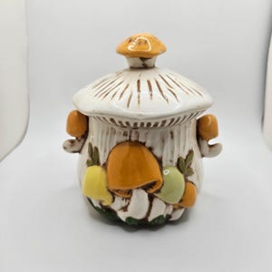 May include: A ceramic mushroom-shaped container with a lid. The container is white with brown vertical lines and orange, yellow, and green mushroom accents. The lid has brown lines radiating from the center. Two small orange mushroom handles are on the sides.
