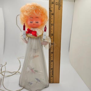 May include: A vintage Christmas tree topper in the shape of an angel. The angel has a peach-colored head with closed eyes and a red bow. It holds two red light bulbs and wears a white dress. A wooden ruler is next to the angel.