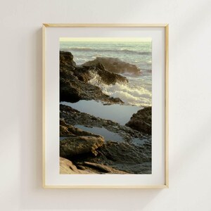 May include: A framed print featuring a coastal scene. The image captures the ocean waves crashing against dark, textured rocks. The water is a mix of blues and greens, with the sky in soft yellow tones.